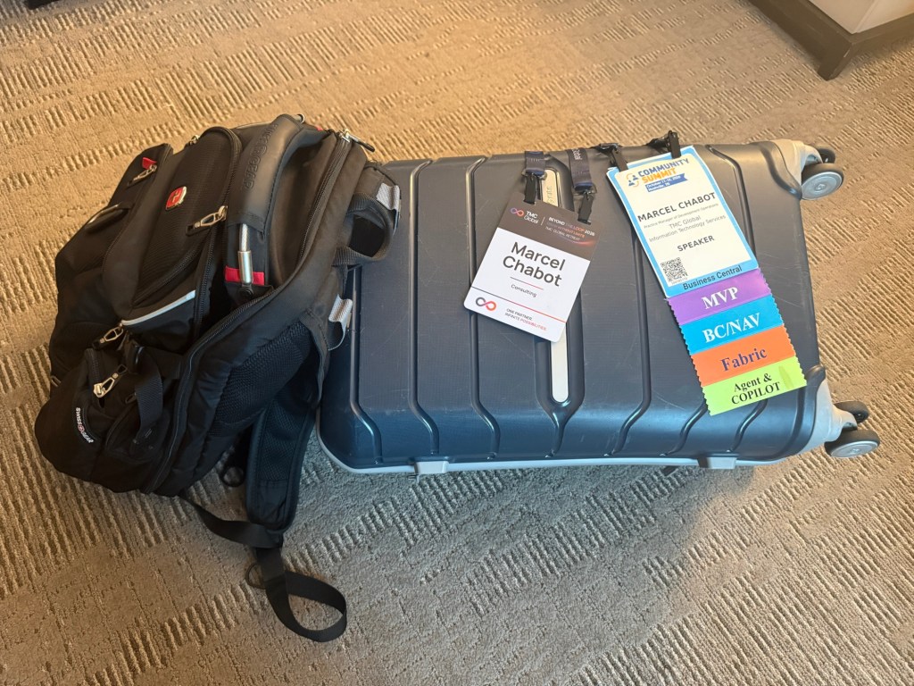 A black backpack and a gray suitcase positioned on a carpet, with name tags attached to the suitcase indicating a speaker at a conference.